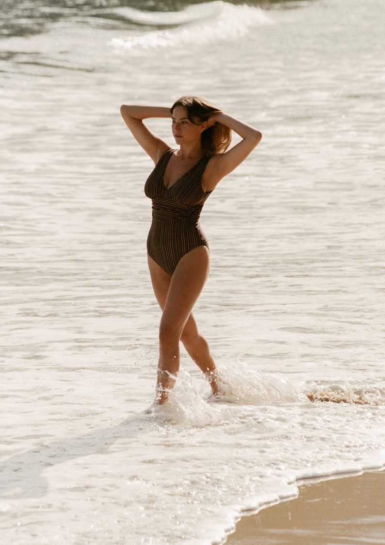 Bokumaia model walking through ocean surf wearing black and brown stripe one-piece swimsuit with sculpted waist
